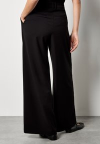 Pantaloni neri a gamba larga realizzati in morbido tessuto, con tasche laterali, vita alta e orlo pulito. Indossati con scarpe nere basse.