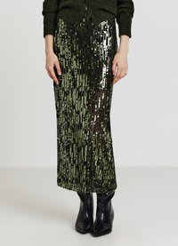 Jupe noire à sequins avec des accents verts verticaux, tissu transparent et silhouette ajustée. Associée à des bottines noires à bout pointu.