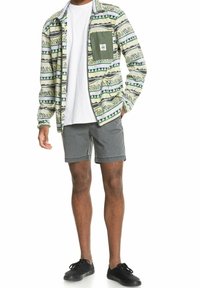 Camisa de manga larga con un patrón en verde, beige y negro, con un bolsillo en el pecho, combinada con pantalones cortos grises y zapatos negros.