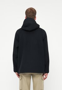 Mees mustas kapuutsiga jope ja beežides cargo-pükstes, seismas seljaga lihtsa valge tausta poole.