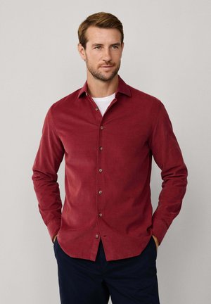 Camisa de botones color marrón con cuello clásico, mangas largas y una textura suave. Presenta botones y un dobladillo curvado. Se lleva con una camiseta blanca debajo.