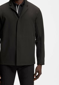  svart strukturerad jacka med slagkrage, sl�ät yta, långa ärmar och sidofickor, bärs över en svart polotröja med vita detaljer.