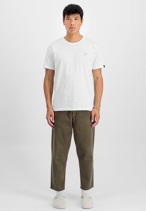 T-shirt en coton blanc avec un col rond et un petit logo doré, associé à un pantalon vert olive et des chaussures slip-on grises claires.