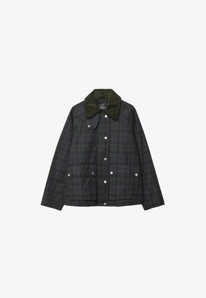 Kvinde iført en mørkegrøn jakke, sennepsgul knappet cardigan, vide mørke jeans, brun bælte og brune sko, står op ad en grå væg.