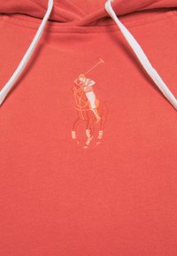 Korallfarbene Kapuzenjacke aus weichem Material mit einem schlichten, gestickten Polo-Spieler-Logo in Orangetönen. Weiße Kordelzüge.