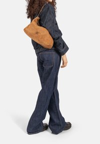 Braune Wildlederhandtasche mit geschwungener Form, integriertem Griff und dezenten Metallakzenten. Kombiniert mit einer Jeansjacke und Jeans mit weitem Bein.