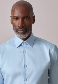 Šviesiai mėlyna marškinėlių tipo palaidinė su smailiu apykakliu, lygiu tekstūra ir perlamutriniais mygtukais, pasižyminti aptakiu siluetu ir be raštų.