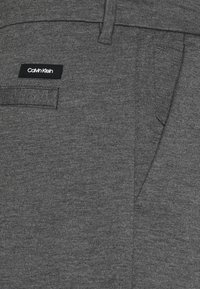 Pantaloni grigi Calvin Klein con una texture liscia, tasche laterali e un'etichetta con il marchio in vita. Design semplice e moderno.