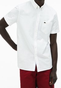 Biała koszula na krótki rękaw z guzikiem, wykonana z bawełny, z kieszonką na piersi i małym zielonym logo. Połączona z czerwonymi spodniami.