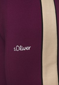 Burgundijske tajice s bež vertikalnom prugom i crnim detaljima. Ime branda "s.Oliver" izvezeno je bijelo na bočnoj strani.