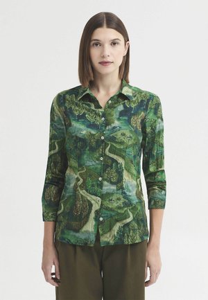 Femme aux cheveux bruns lisses portant une chemise verte à boutons avec un motif de paysage détaillé et un pantalon marron sur un fond uni.
