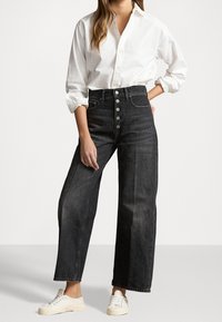 Mujer vistiendo una camisa blanca de botones metida dentro de unos pantalones vaqueros de pierna ancha alta, negros, con botones en la parte delantera y zapatillas blancas, en una pose de pie.