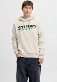 Bež hoodica s prednjim džepom, s natpisom "STUDIO" u debelom crnom fontu i "Jack & Jones" u zelenoj kurvnom pismu. Nosiva s tamnim trapericama.