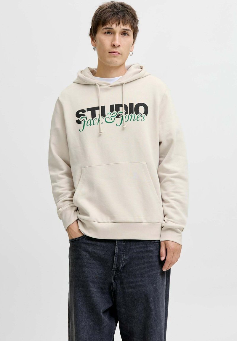Bež hoodica s prednjim džepom, s natpisom "STUDIO" u debelom crnom fontu i "Jack & Jones" u zelenoj kurvnom pismu. Nosiva s tamnim trapericama.