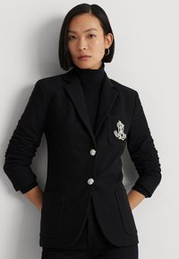 Zwarte blazer met een Tailored ontwerp, zilveren knoopaccenten en een borstzak met een geborduurd logo. Textuurstof en getailleerde silhouette.