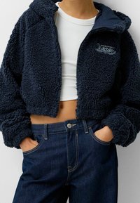 Mörkblå fleecetröja med huva, kort design, texturerad yta och broderad "Oregon"-logotyp. Kombination med mörka denimjeans.