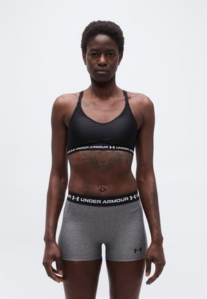 Atletisk kvinde iført sort Under Armour sports-bh og grå kompressionsshorts står vendt mod kameraet foran en ensfarvet baggrund.