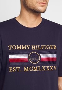 Námornícky bavlnený tričko s vyšitým zlatým textom "TOMMY HILFIGER" a logom. Obsahuje pruhovaný vzor v červenej, bielej a námorníckej modrej pod textom.