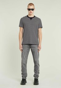 Czarny i szary pasiasty t-shirt z krótkim rękawem oraz dekoltem w serek, zestawiony z jasnoszarymi dżinsami i czarnymi trampkami. Model nosi ciemne okulary przeciwsłoneczne.