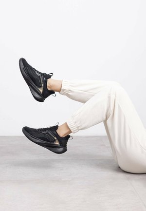 Zapatillas deportivas negras con parte superior de malla, acentos dorados y suela negra. Combinadas con pantalones de chándal de color claro, piernas levantadas contra un fondo neutro.