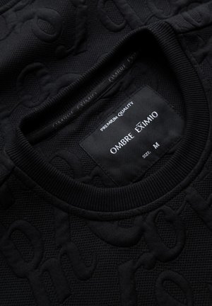 Schwarzer strukturierter Pullover mit geripptem Halsauschnitt und geprägtem Muster. Das Etikett trägt "Ombre Eximio", Größe M, mit dem Slogan "Premium Quality".