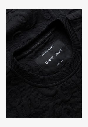 Suéter negro texturizado con un cuello de canalé, que presenta un patrón en relieve. La etiqueta dice "Ombre Eximio", talla M, con el lema "Calidad Premium".