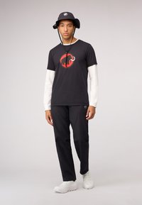 Zwarte t-shirt met rood olifantenlogo, gelaagd met een witte lange mouw, gecombineerd met zwarte broek en witte sneakers, afgewerkt met een zwarte bucket hat.