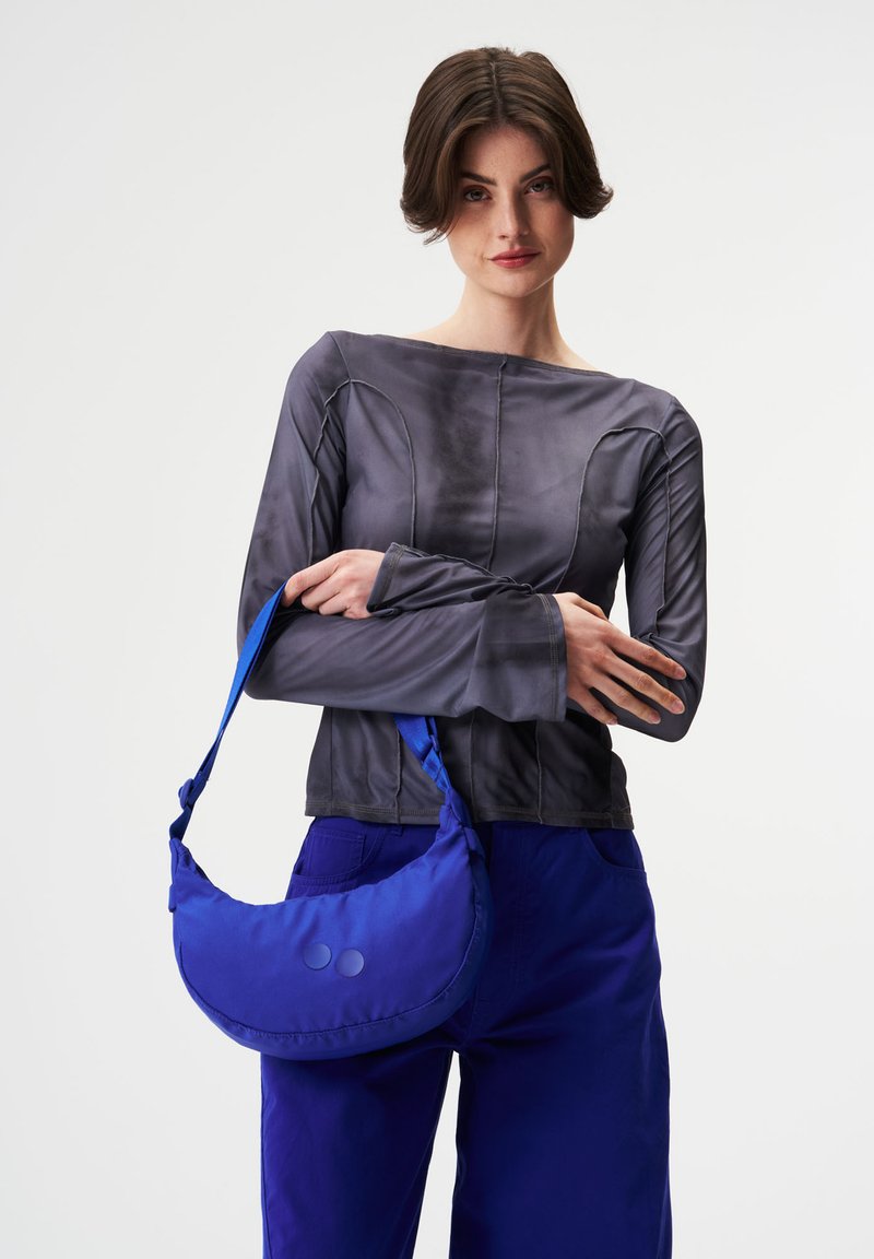 Blaue Stoff-Schultertasche mit zwei runden Akzenten, kombiniert mit einem grauen Langarmoberteil mit strukturierten Nähten und einem quadratischen Ausschnitt.