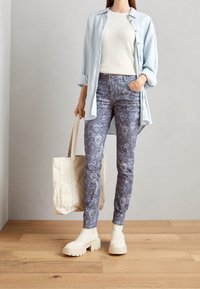 Ljusblå knapp-up skjorta över en krämfärgad topp, tillsammans med mönstrade blå jeans och vita klumpiga plattformsskor. Inkluderar en beige tote-väska.