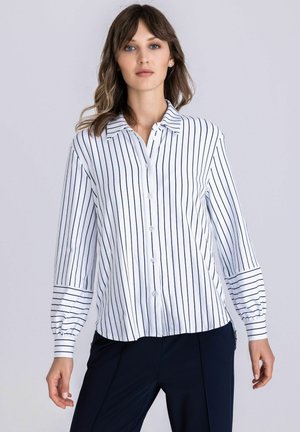 Woman wearing a white button-up blouse with thin vertical black stripes and long puffed sleeves, paired with dark trousers, standing.