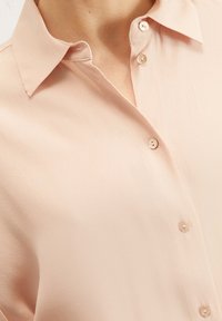 Světle růžová blůza s límečkem, která má hladkou texturu, viditelné detaily knoflíků a volnější střih kolem ramen.