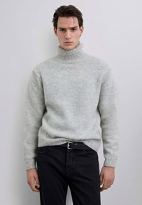 Jersey de cuello alto gris claro con cuello y puños acanalados, con una textura suave y un ajuste relajado, combinado con pantalones vaqueros oscuros.