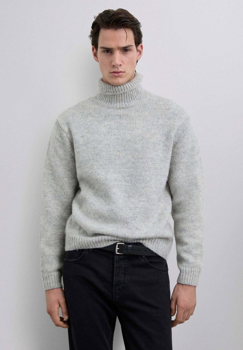 Jersey de cuello alto gris claro con cuello y puños acanalados, con una textura suave y un ajuste relajado, combinado con pantalones vaqueros oscuros.