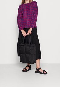 Gestepptes schwarzes Tote-Bag mit dicken Trägern, kombiniert mit einem lila-blau karierten Langarmshirt und einem schwarzen gefalteten Rock. Legere Sandalen runden das Outfit ab.