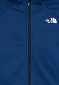 Casaco de fleece azul com fecho frontal, elementos pretos e um logótipo branco no lado esquerdo do peito. Textura suave e design ajustado.