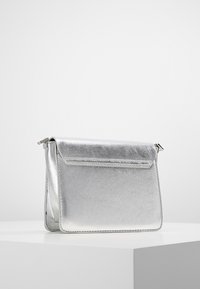 Malá stříbrná metalická kožená kabelka s klopou, položená na bílém podstavci na bílém pozadí.