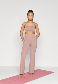 Σετ γυμναστικής σε ανοιχτό ροζ χρώμα, αποτελούμενο από κοντό αθλητικό σουτιέν και ψηλόμεση καμπάνα παντελόνι, φτιαγμένο από υφή υφάσματος, εκτιθέμενο σε ροζ στρώμα.