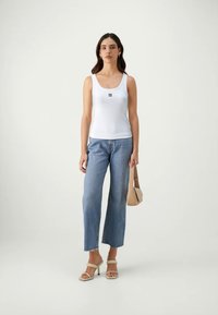 Weißes Tanktop mit kleinem schwarzem Logo, kombiniert mit hellblauen weiten Jeans und beigefarbenen Absatzsandalen. Die Accessoires umfassen eine beigefarbene Handtasche.