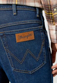 Jeans de mezclilla azul oscuro con un parche de cuero marrón con la marca "Wrangler". Costuras amarillas distintivas y un diseño de bolsillo trasero angular.