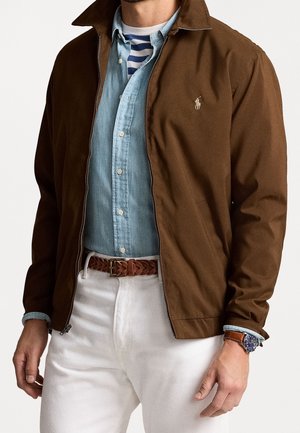 Man wearing brown zip jacket over light blue button shirt and striped t-shirt, white pants, woven brown belt, and wristwatch.