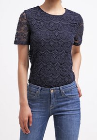 Femme portant un haut en dentelle bleu marine à manches courtes associé à un jean bleu délavé sur fond uni.