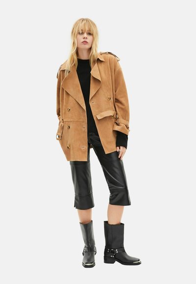 Gabardina de ante color canela con botones cruzados, pantalones cortos de cuero negro y botas negras de tobillo con detalles metálicos.