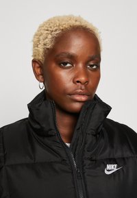 Černá péřová bunda s vysokým límcem, vyrobená z lesklého materiálu, se zapínáním na zip a bílým logem Nike na hrudi. Viditelné detaily texturované izolace.