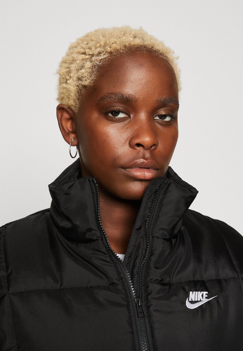 Black puffer jacket with high collar, featuring glossy fabric, zip closure, and a white Nike logo on the chest. Textured insulation detail visible.