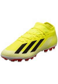 Botín de fútbol amarillo con un upper texturizado, diseño de rayas negras y tacos rojos en la suela. Cuenta con un cuello ceñido tipo calcetín.