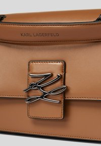 Strukturalna torba ze skóry w kolorze tan z magnetycznym zapięciem. Posiada srebrny akcent z logo, proste linie i widoczne szczegóły szwów.