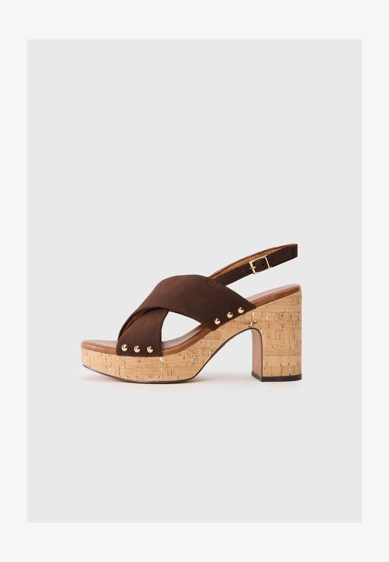 Sandale à talon haut en suede marron avec un talon en liège. Présente un design à brides croisées, une bride de cheville réglable et des accents en clous dorés sur le côté.