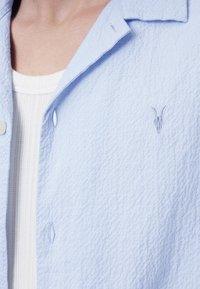 Lichtblauw gestructureerd overhemd met kraag, voorzien van een geborduurde geiten schedel logo op de borst. Daaronder is een witte geribde top.