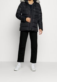 Chaqueta de plumas negra con capucha con borde de piel, con diseño acolchado, cremallera frontal y botones de presión. Llevada sobre un top azul marino y pantalones negros.