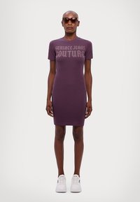 Fialové upnuté šaty s krátkými rukávy a kulatým výstřihem, s potiskem "Versace Jeans Couture" v světle šedé barvě napříč hrudníkem. V kombinaci s bílými teniskami.